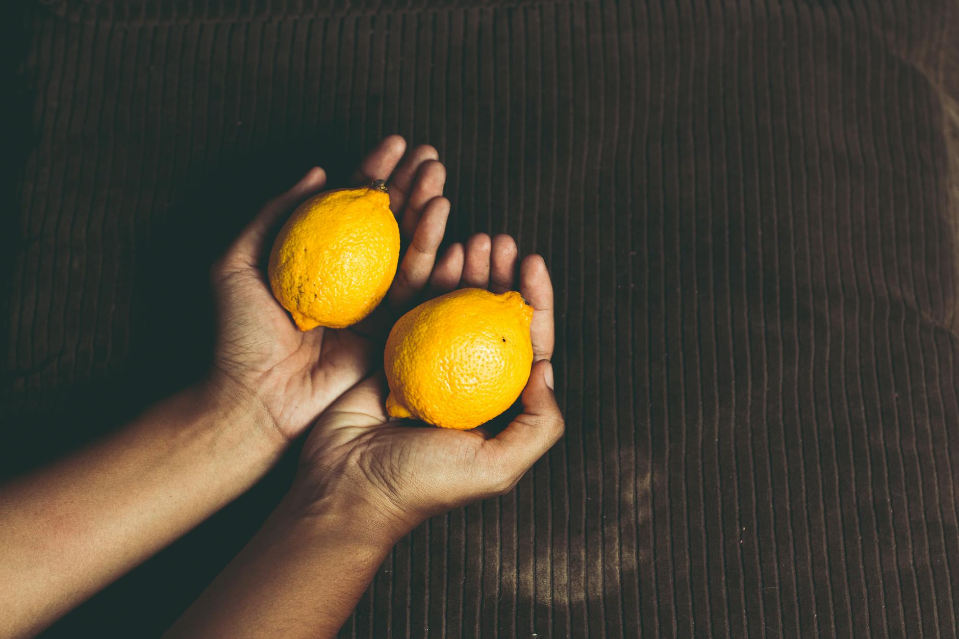 Close-up of two fresh lemons held in cupped hands on a brown surface, representing the lemon clitoral vibrator design philosophy.